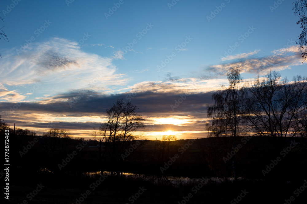 Fototapeta premium Stunning sunset over a tranquil landscape. Silhouetted trees frame the vibrant sky, reflecting in the calm water. A peaceful, golden hour scene