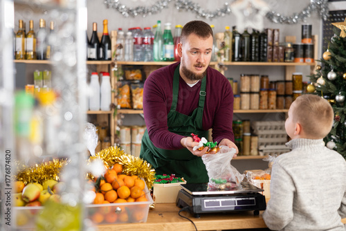 Kind salesman assisting little boy with holiday candies purchase in decorated store