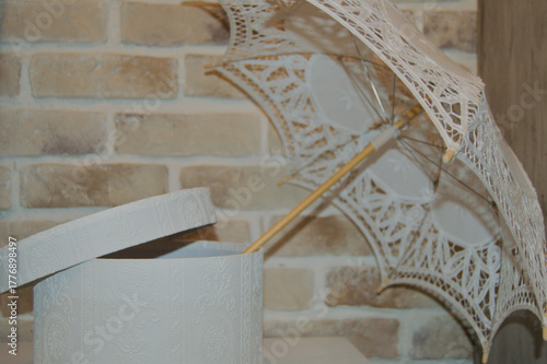 Decorative lace parasol and ornate white gift box with embossed texture, arranged on a light surface in front of a rustic brick wall. The image conveys a sense of elegance, vintage charm