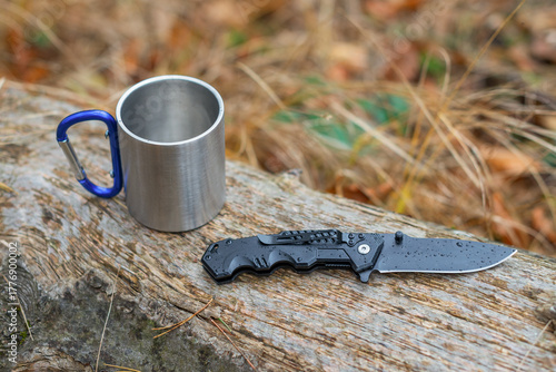 Outdoor camping gear on a rainy day with a folding knife and a metal mug on a wooden log