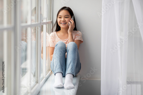 Free Download A young girl sits on a windowsill, smiling and chatting on her phone. Sunlight fills the room, creating a cheerful and warm atmosphere. She wears a striped shirt and denim jeans. - GFXLoad