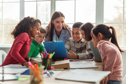 Joyful school children engage with their teacher in a dynamic learning session at their desks. They explore study materials together in a bright, inviting classroom setting.