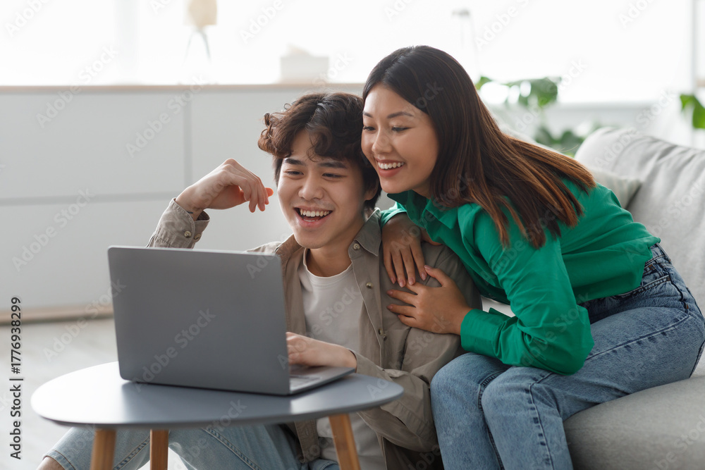Naklejka premium A young Asian couple dressed casually sits together on a sofa, happily watching a movie on a laptop. They are engaged in an online meeting, enjoying their shared time at home.