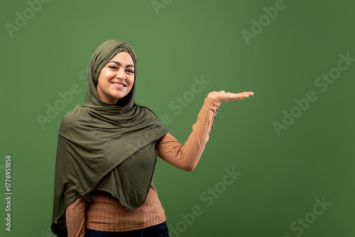 A cheerful arab lady wearing a hijab stands confidently on a dark green background, pointing to the side with her open palm as if presenting something to the audience. She looks friendly and inviting.