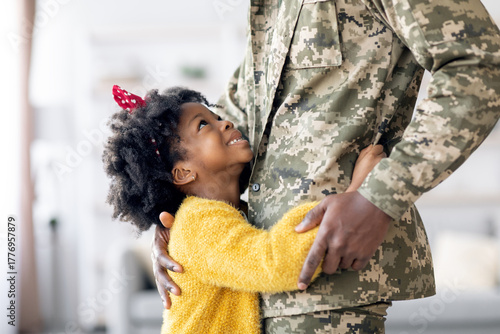 Foto In a warm and cozy living room, a young girl wearing a yellow sweater joyfully hugs a service member in military uniform