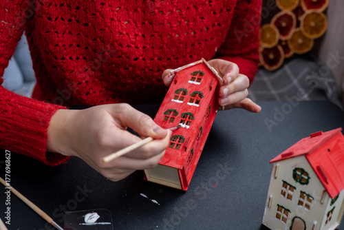 Sustainableminded Christmas cozy atmospheric festive decor in modern scandinavian home handmade decoration. Unrecognizable woman in red sweater painting craft wooden houses. Seasonal activity for kids