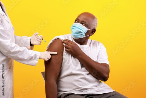 A healthcare professional is giving a COVID vaccine shot to an elderly black male patient in a bright orange studio background. The man is wearing a medical mask and appears calm.