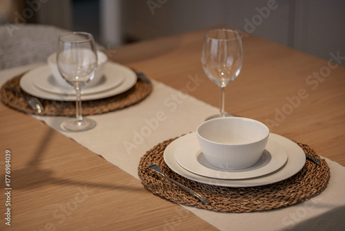 Elegant table setting for two. Minimalist white dinnerware, wine glasses, and wicker placemats on a wooden table. Dining concept.