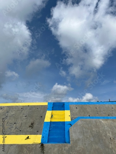 Low-angle photo of yellow and blue painted concrete surface against bright sky and white clouds.
