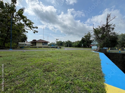 Tropical field with blue and yellow painted canal beside trees and cloudy afternoon sky.