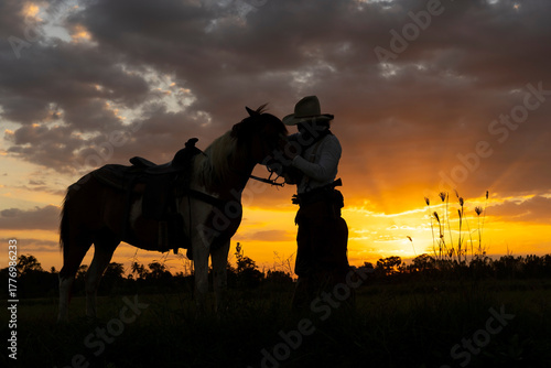 Silhouette of a cowboy standing beside his horse in the golden light of sunset on the western plains. Peaceful and cinematic moment capturing freedom and western lifestyle.