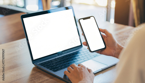 A close-up of a person using a laptop and holding a smartphone, both with blank white screens, on a wooden table. Perfect for showing work, mockups, or app use.