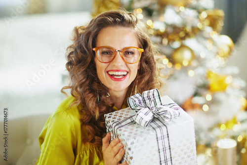 A cheerful woman with curly brown hair and glasses holds a gift box, smiling warmly in a close-up against a softly blurred Christmas tree adorned with golden lights.