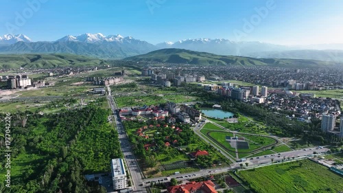 Wide aerial view of a modern area of the Bishkek city with apartment buildings, a central park, roads, and lush green foothills leading to the snow-capped Tian Shan mountains