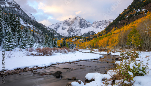 The iconic snow-capped Maroon Bells mountains overlook a snowy meadow and stream. Vibrant yellow aspen trees line the valley, showing a transition from autumn to winter.