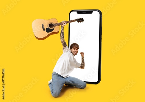 Big smartphone and young tattooed man with guitar on yellow background