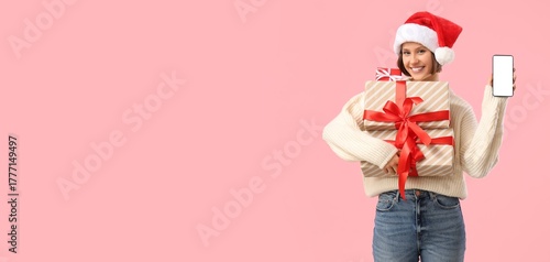 Young woman in Santa hat holding gift boxes and mobile phone on pink backgrou...