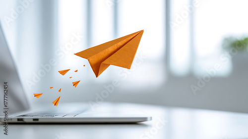 Orange paper airplane taking off from a laptop with smaller planes following behind it on a white table