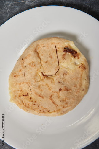A close-up photo of a golden, cheese-filled pupusa on a white plate against a black-grey marble background.