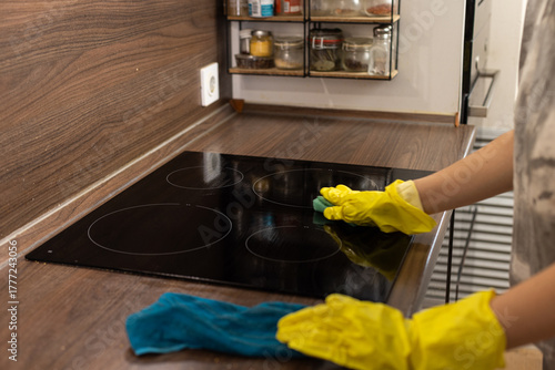 A person wearing yellow gloves scrubs a soapy cooktop with a sponge, removing grease and grime from the kitchen surface.