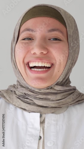 Vertical video. Cheerful young muslim healthcare professional in hijab smiling warmly in a studio headshot, representing diversity and care