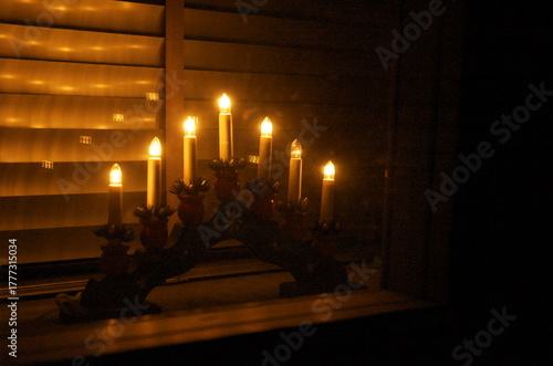 A glowing candle arch sits softly in a window, its warm light reflecting across wooden blinds. A quiet winter moment, evoking comfort, tradition, and the gentle calm of December nights.