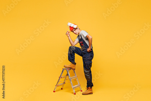 Full body young sad employee laborer handyman man wear helmet overall stand on stepladder use paint roller isolated on plain yellow background Instrument renovation apartment room Repair home concept