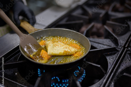 Chef cooking orange butter sauce for Crepes Suzette on gas stove, dessert caramelization process in professional kitchen environment.