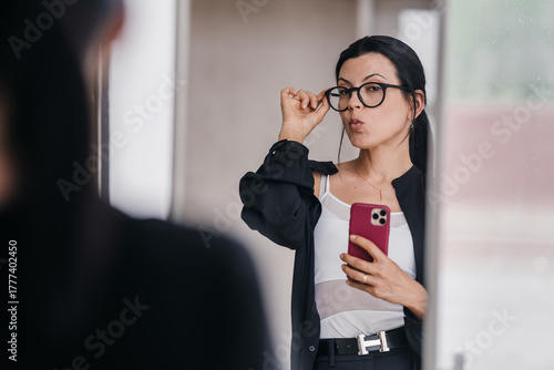 A confident woman captures her reflection while adjusting eyeglasses, exuding poise and charisma in a casual setting.