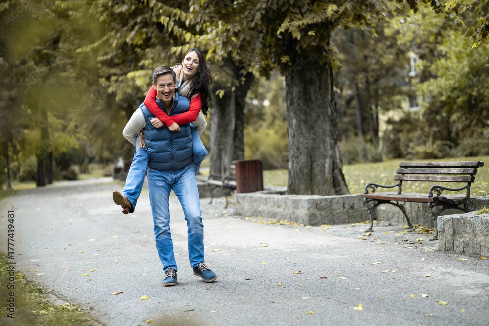 Fototapeta premium The playful couple enjoys a joyful piggyback ride in the park, displaying their love and happiness; a perfect moment captured amidst the beautiful autumn foliage and scenery.
