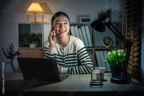 happy woman talking with smartphone while working with laptop at night
