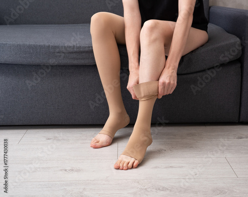 Unrecognizable woman dressing legs in elastic compression hosiery at home interior promoting vascular health, comfort and medical prevention therapy