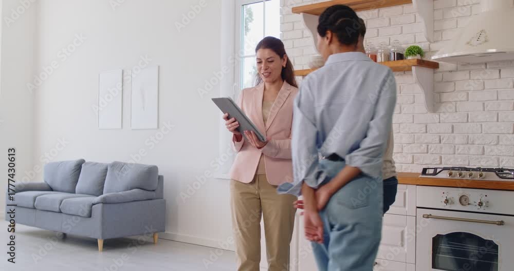 Couple meeting with real estate agent for home apartment tour. Happy realtor greeting woman and man with handshake, showing property plan or agreement on tablet and room interior, talking to clients
