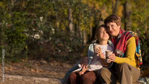 family grandmother and  child granddaughter  hugging and drinking tea from a thermos on the riverbank on a sunny autumn day.  Family leisure with kids at autumn.