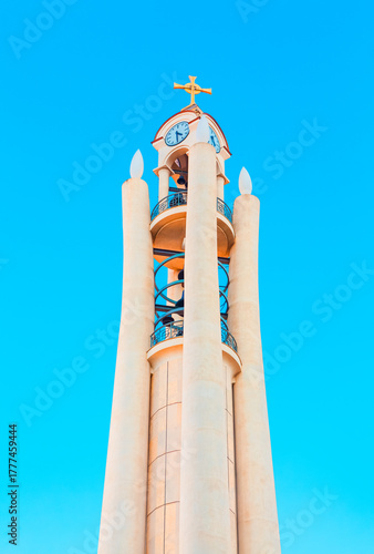 Modern bell tower of the Resurrection of Christ Orthodox Cathedral in Tirana, Albania. Its unique design features a clock face and a spiraling staircase, topped by golden cross against clear blue sky