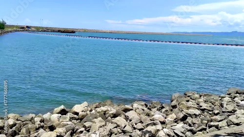 Calm blue water laps against a rocky shore with a floating barrier in the distance under a clear sky
