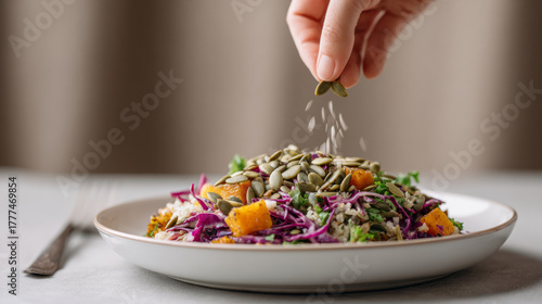 Close-up of hand sprinkling pumpkin seeds on colorful healthy salad with purple cabbage and roasted vegetables on white plate