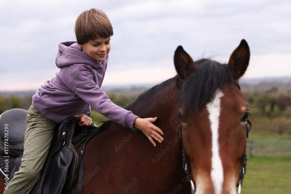 Fototapeta premium Equine assisted therapy. Little boy stroking horse while riding outdoors. Lovely domesticated pet