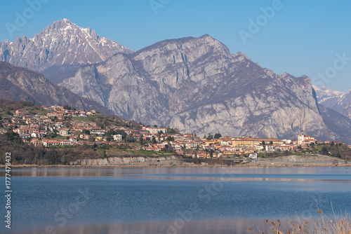 Fototapeta Naklejka Na Ścianę i Meble -  Serene landscape featuring a tranquil lake with a small town nestled on a hillside, beneath majestic, snow-capped mountains