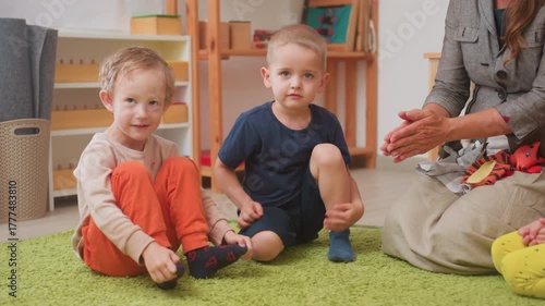 Preschool scene with two boys seated on rug while adult hands demonstrate object off frame, children split attention, curious expressions, classroom shelves and toys visible, casual learning