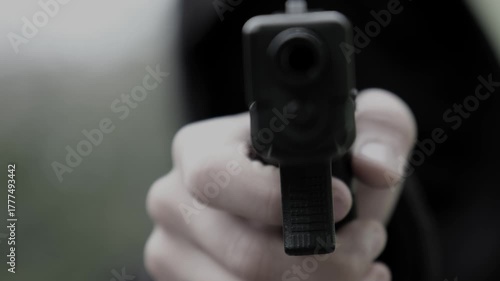An unrecognizable killer or man in a hood and mask points a gun and shoots directly at the camera or the victim. Close-up front view.