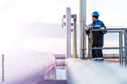 A male worker in a blue hard hat and jacket stands on a platform, watching a system of industrial oil pipelines, the pipes of which extend into the distance under a clear sky. copy space