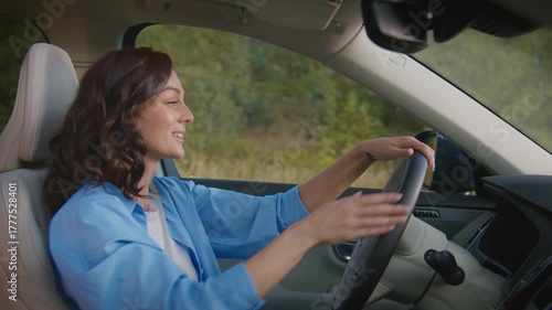 Caucasian woman with wavy hair driving car and talking hands free using built-in speakers. Female communicating safely while keeping focus on road. Relaxed conversation during commute.