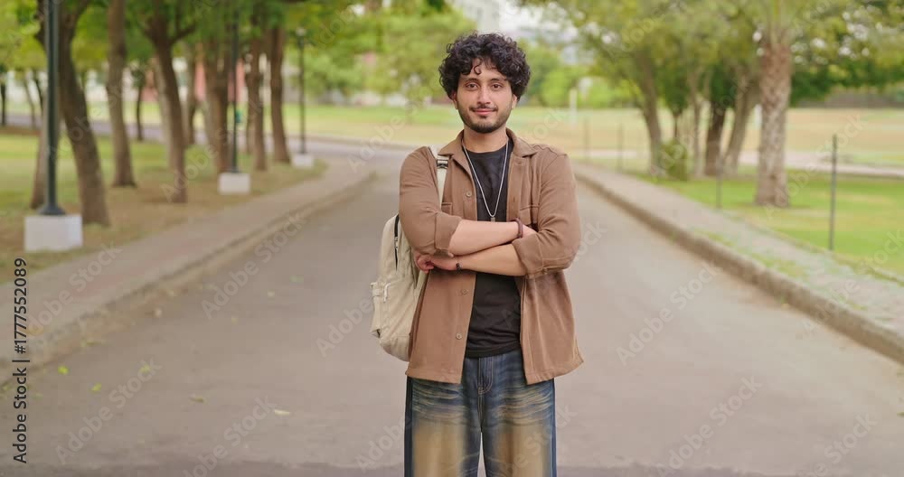 Indian happy young adult gen z man male look camera cross arm standing outdoor area. Smart cool proud pupil boy guy wear bag fold hand staring cam do casual pose enjoy new day outside place