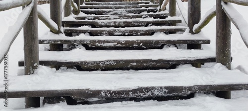 A wooden staircase. The flat wooden steps are covered in white snow. Footprints are visible in the snow. The steps are arranged horizontally, one above the other, along the edges of the railing.