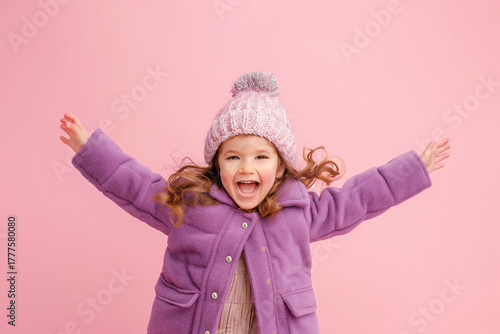 Joyful girl wearing purple coat and pink hat celebrating with arms wide open against pink background