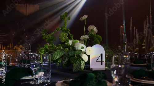Luxury wedding reception. Festive table setting with flower arrangement, number 4 for guests on black tablecloth, green napkins, candles on backdrop, soft moving searchlight rays in hall restaurant.