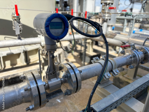 Ultrasonic flow meter mounted on industrial pipeline with digital display. Close-up of metallic pipe, flanges, bolts and connected cable. Background shows additional valves and piping.