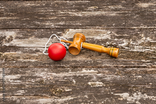 Single traditional Japanese cup-and-ball toy (kendama) with red ball and string, displayed on a weathered rustic wooden table. Outdoor. Symbolizes traditional games, skill, and Japanese culture.