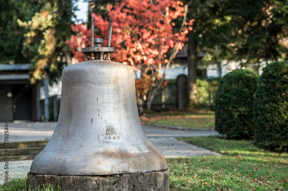 Obraz premium Abgehängte Kirchenglocke mit Jahreszahl 1949 auf Steinsockel vor Baum mit Herbstlaub und Hecke beim Friedhof in Reutlingen-Mittelstadt